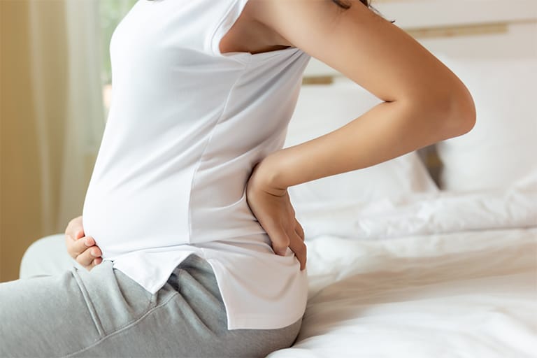Pregnant woman sitting on a bed holding her back in discomfort.